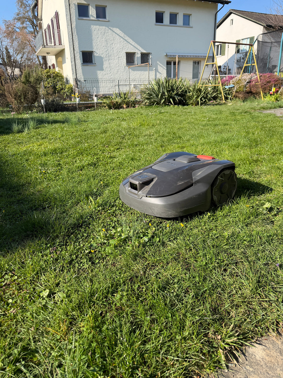 Husqvarna Automower 308V Mähroboter mit Vision-Kameratechnologie bei einer Testinstallation auf einer grünen Rasenfläche vor einem Wohnhaus.