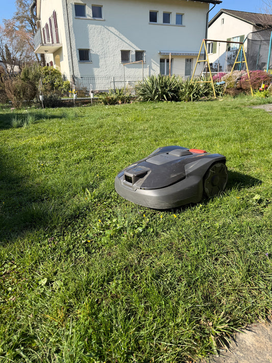 Husqvarna Automower 308V Mähroboter mit Vision-Kameratechnologie bei einer Testinstallation auf einer grünen Rasenfläche vor einem Wohnhaus.